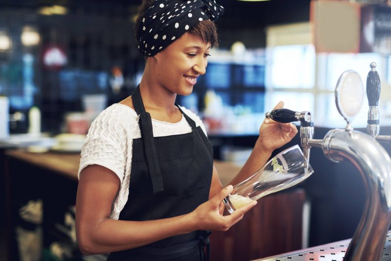 Young attractive female African American bartender pouring a glass of draft beer from the keg dispensing it from a tap behind the counter with a smile